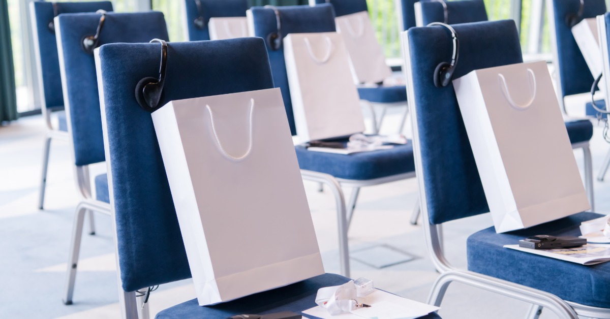 A business seminar set up with chairs in rows. Each chair has a white gift bag full of promotional items.