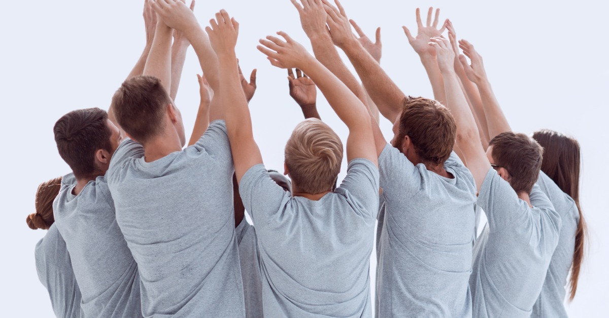 A group of employees standing together in a circle with their arms up as they cheer. They're wearing matching gray shirts.