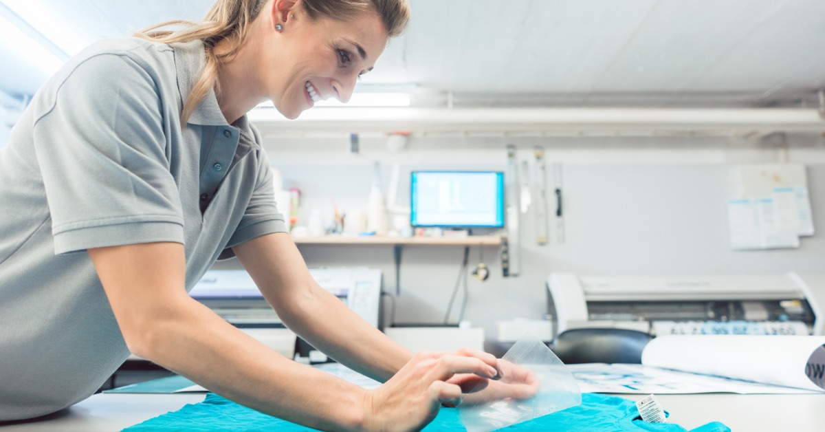 A custom shirt designer smiles as she places a screen print on a bright teal shirt laying on the counter.