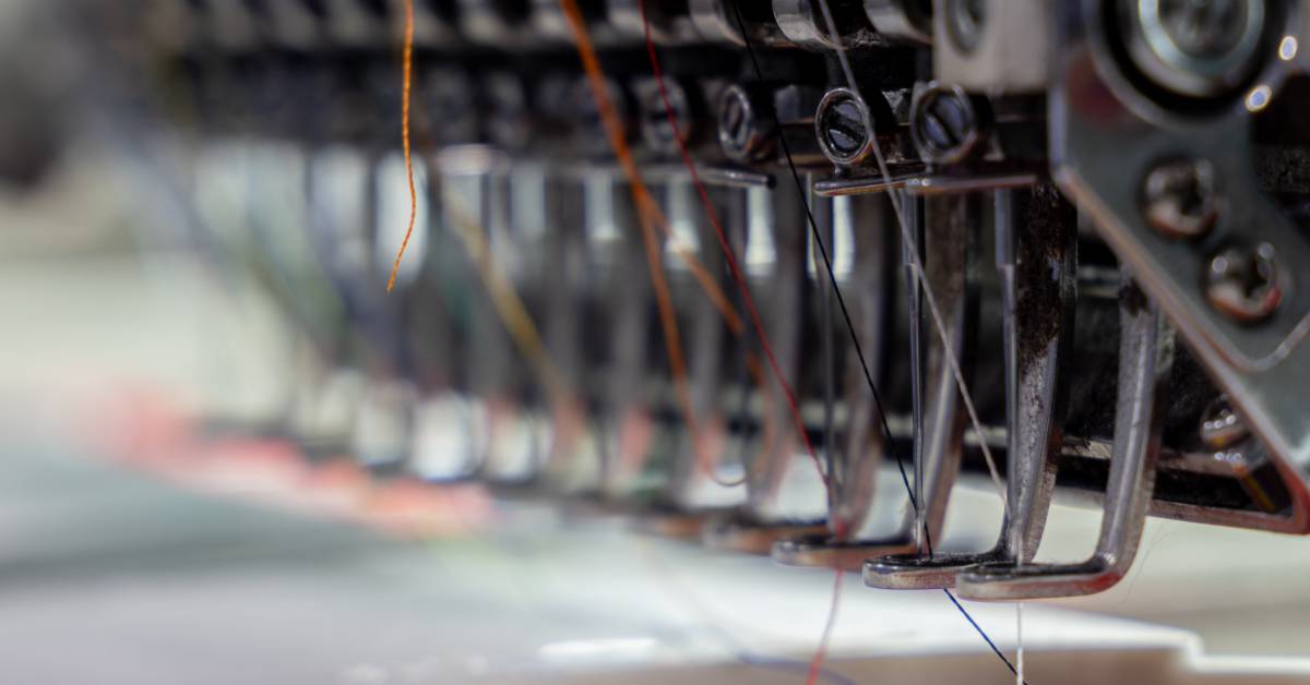 A close-up of a commercial embroidery machine needle and thread part. The threads are different colors.