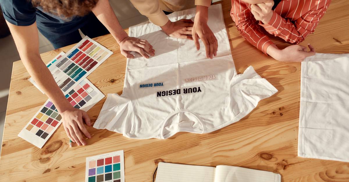 A wooden table where three designers stand at the edge to review a custom logo T-shirt and color samples for a brand.