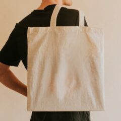 A person holds a canvas tote bag without a design on it The person wears a black T-shirt and faces the wall