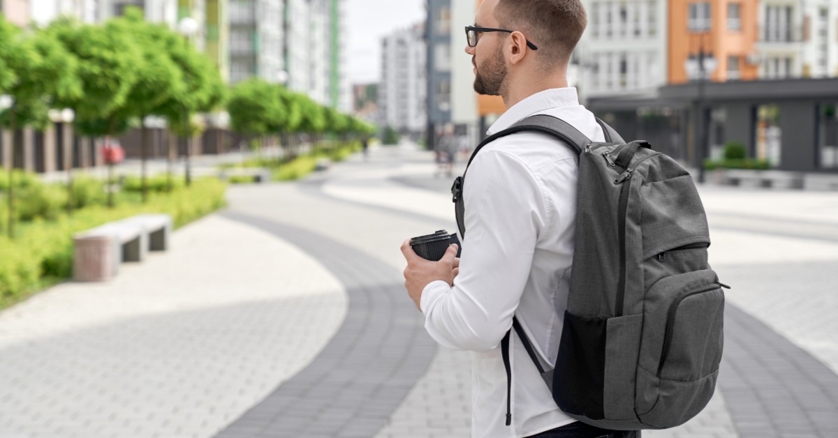An employee wears a company backpack as he walks around outside in business casual attire. He holds a coffee.
