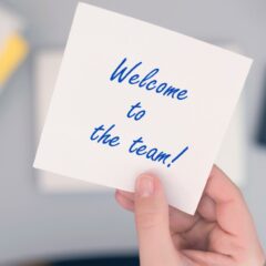 A new employee sits at their desk with a white note that says Welcome to the team in blue writing