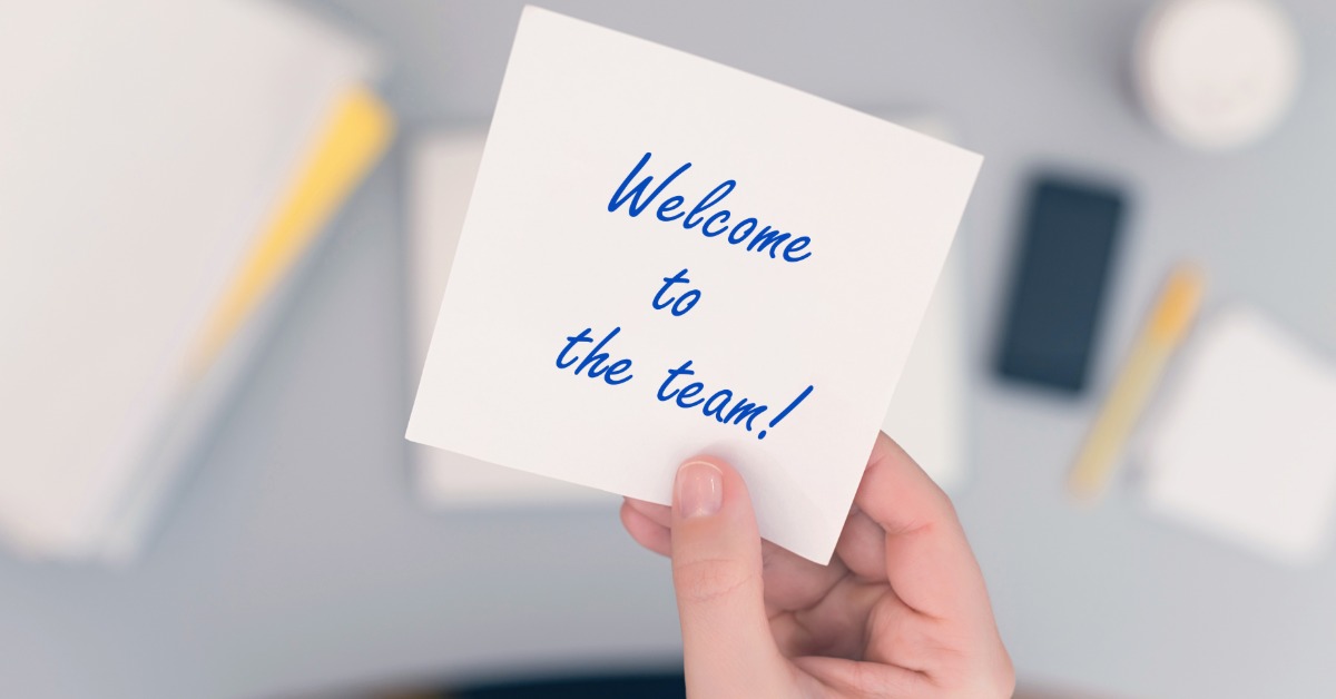 A new employee sits at their desk with a white note that says "Welcome to the team!" in blue writing.