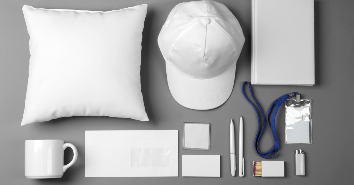 A collection of white, unbranded trade show giveaway items, such as a pillow, hat, and pens, sits on a gray surface.