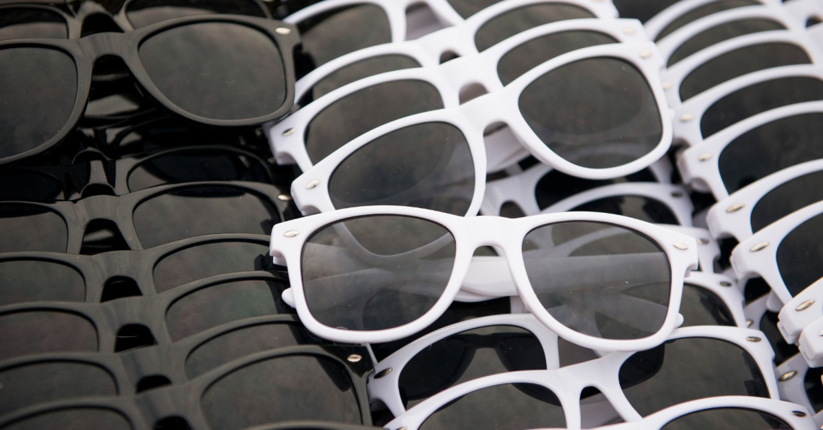 A table with bulk sunglasses lined up ready for customization. There are black options and white options.
