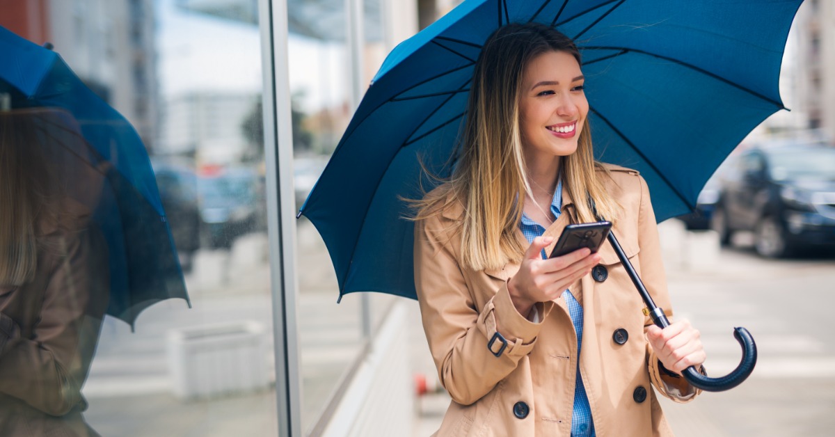 A young business woman walks outside in a trench coat as she holds her phone in one hand and an umbrella the another.