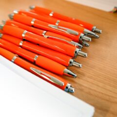 A collection of orange ink pens sit on a desk next to a manila folder and papers The pens need custom logos