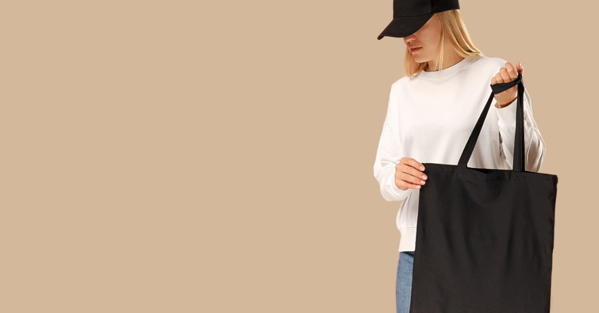 A female model with blonde hair stands against a peach-colored background. She wears a black hat and holds a tote bag.