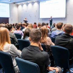 A corporate conference with guests seated in cloth chairs in front of the stage and a guest speaker presenting