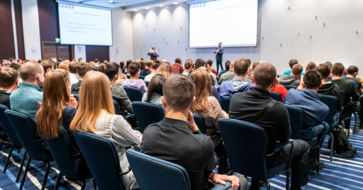 A corporate conference with guests seated in cloth chairs in front of the stage and a guest speaker presenting.