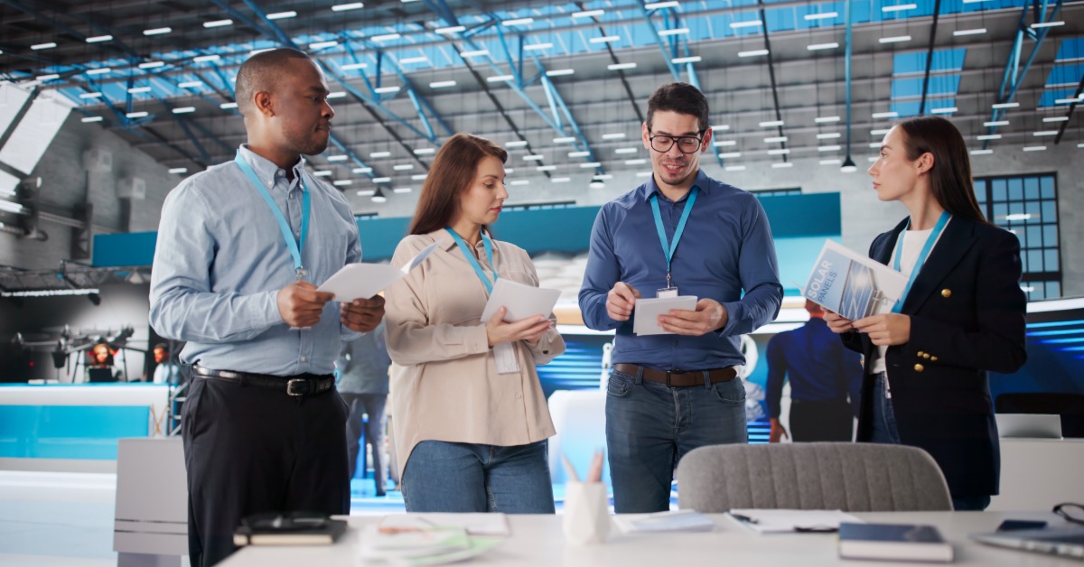A group of four professionals stand at the trade show booth with papers in their hands and lanyards around their necks.