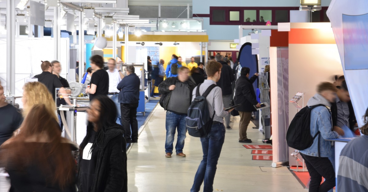 A busy trade show with people walking around. There is a row of trade show booths with people in backpacks.
