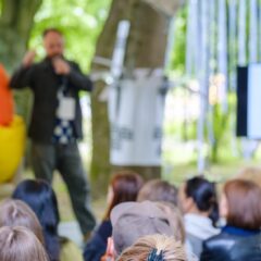 An outdoor business event with a guest speaker on stage talking into a microphone and guests seated