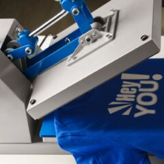 A garment printing press sits on a table with a blue T-shirt placed in the press The graphic is white and says hey you