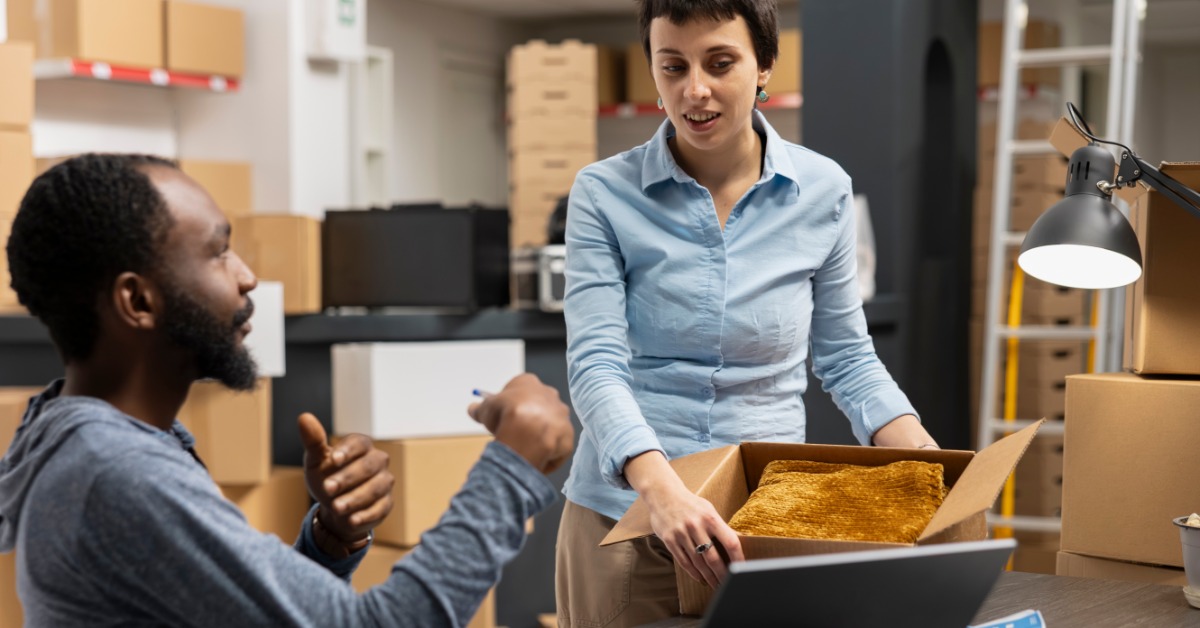 Two team members in the office at the laptop prepare an onboarding kit for a new team member. The box has tissue paper.