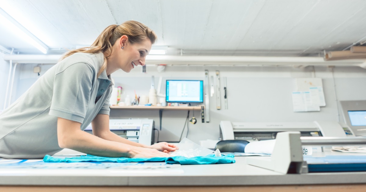 A screen printing professional stands over the work table in the shop. She lines up a T-shirt for printing.