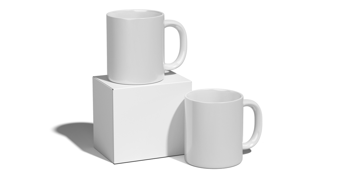 Two blank, white coffee mugs sit on a white background. One coffee mug sits on a plain cardboard box.