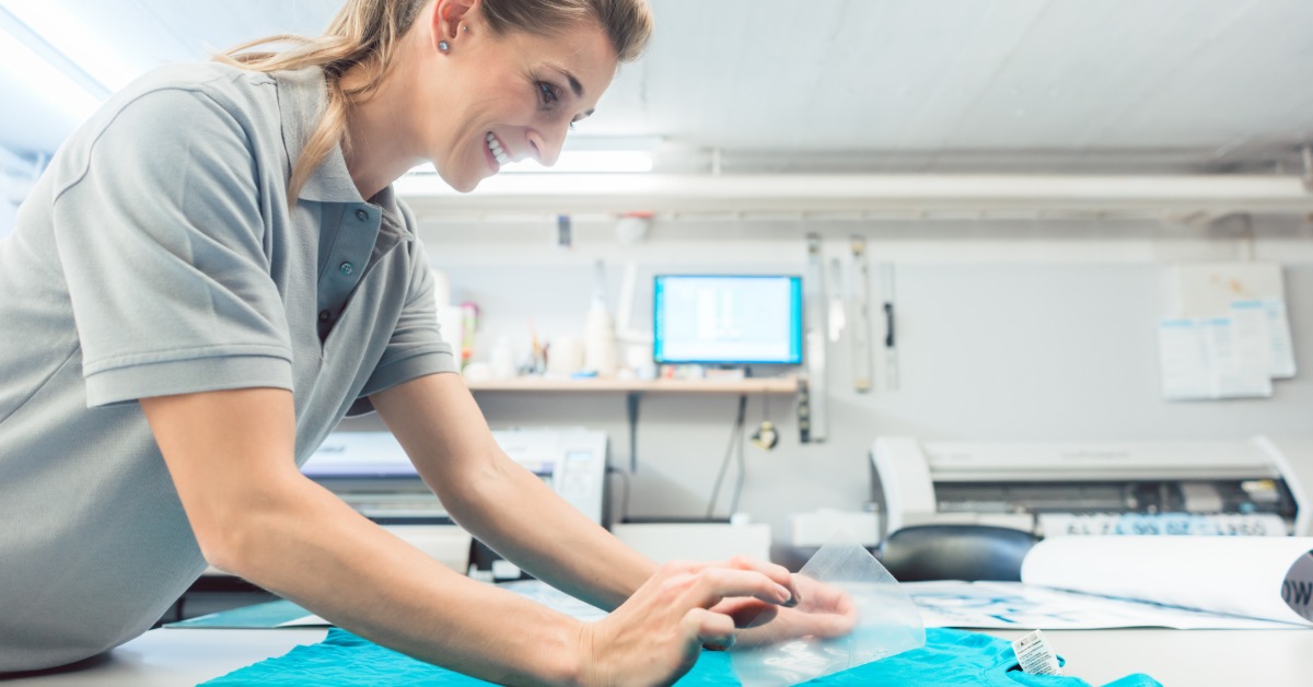 A promotional specialist works at a table on customizing T-shirts for a business. She smiles and the shirt is teal.