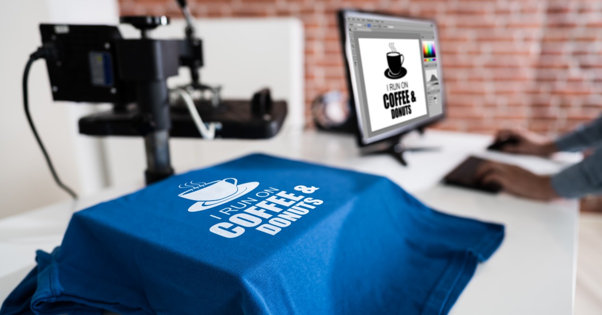 A royal blue T-shirt sits on a heat press ready for customization. Next to the press is a computer with the design.