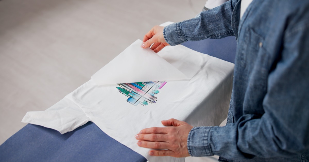 A printing professional stands over a workstation as they peel an iron-on decal off a plain white T-shirt.