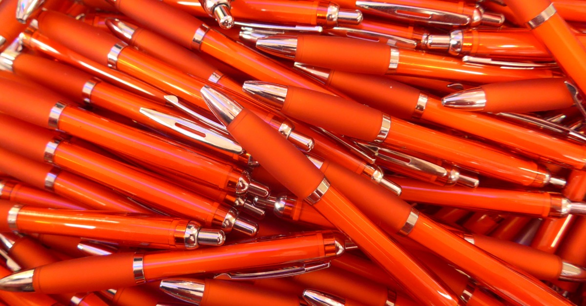 A close-up of a pile of red ink pens. They all have a stainless steel trim around the base and click button.